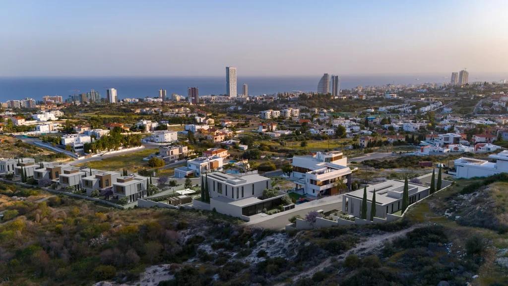 Aerial view of a coastal city with modern houses in the foreground, high-rise buildings near the shoreline, and the sea visible in the background under a clear sky.