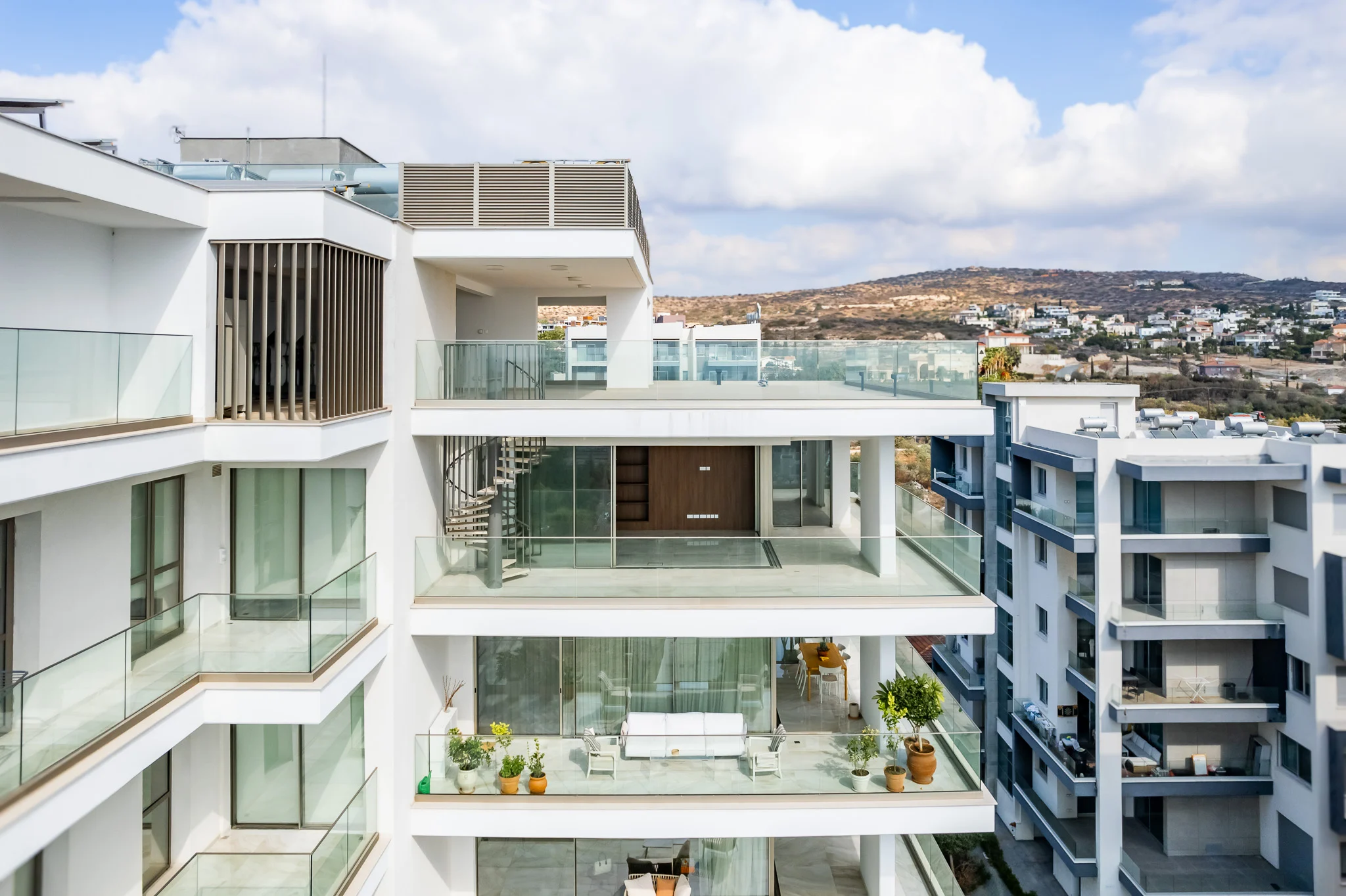 A modern white apartment building with large glass balconies and potted plants, overlooking a hilly landscape with more buildings in the background under a partly cloudy sky.