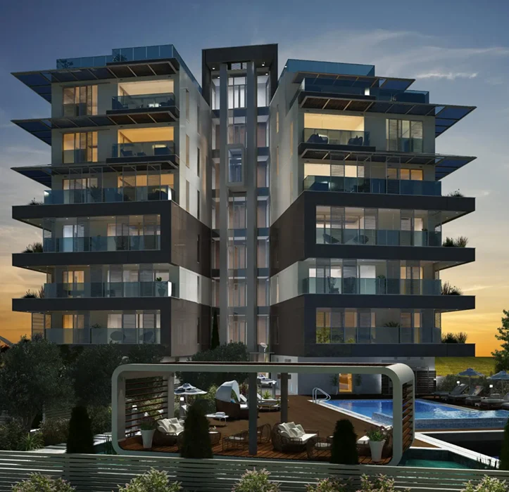 A modern, multi-story apartment building with large glass balconies, illuminated at dusk. In front, there is a stylish outdoor lounge area and a swimming pool, surrounded by greenery and seating.