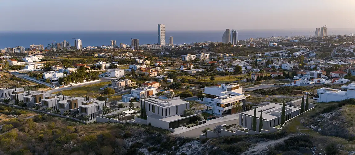 Modern white villas with large windows and balconies are lined up along a quiet street at dusk, surrounded by landscaped greenery and hills in the background. Several cars are parked by the entrance.