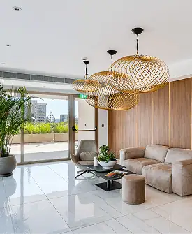 Modern living room with large windows, a beige sectional sofa, wooden accent wall, potted plant, marble floor, and three woven pendant lights. Natural light fills the space, with a view of a balcony and greenery outside.